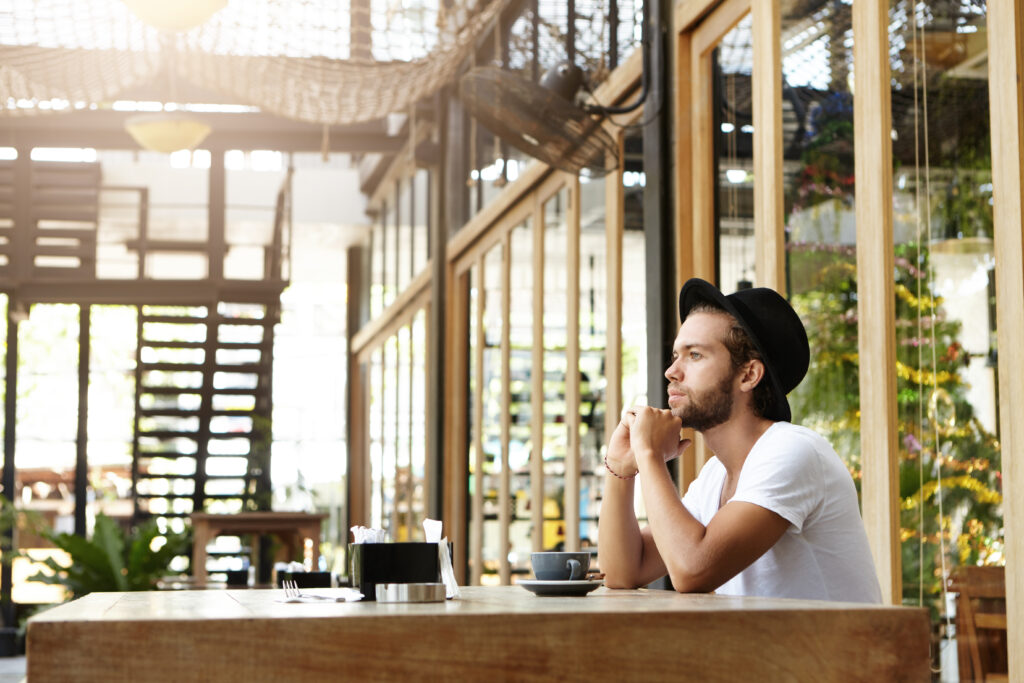 Handsome young writer with thick beard wearing black hat looking pensive and absorbed in his thoughts, leaning on his elbows while thinking on plot of his new book over coffee at sidewalk cafe